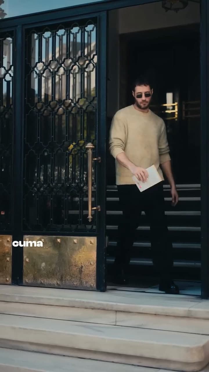 A man in a cream knit stepping out of a wrought-iron doorway.