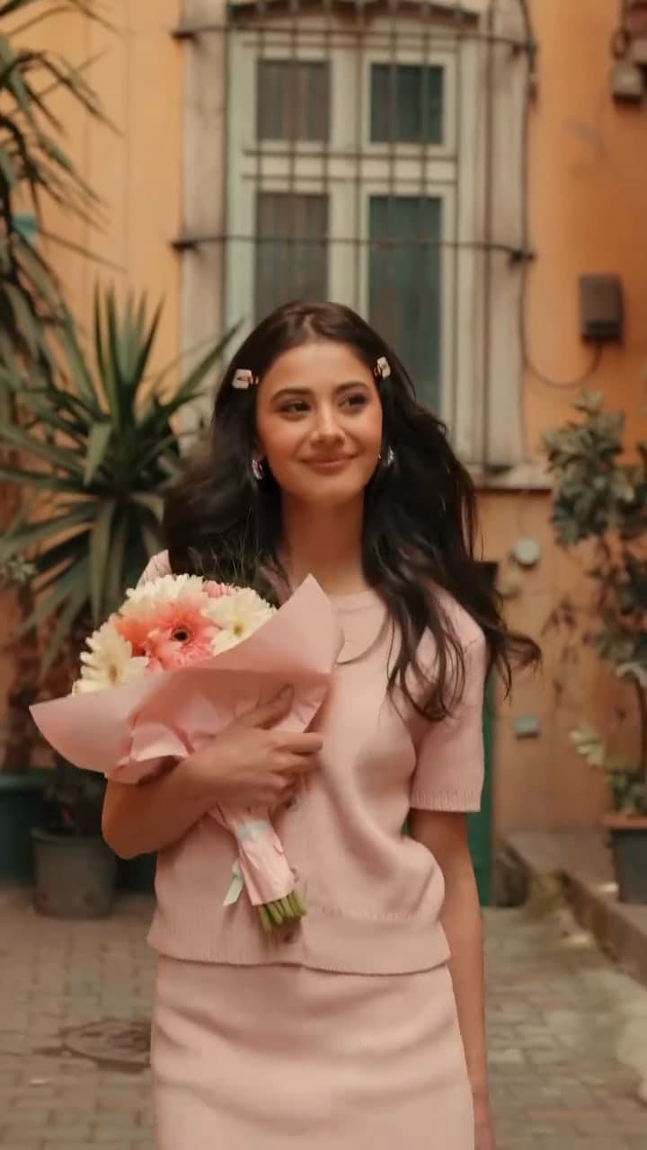 Helin Kandemir in a pink knit set holding a bouquet in a sunlit courtyard.