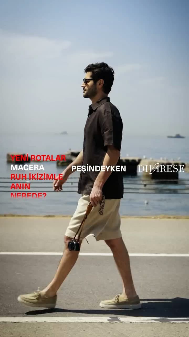 A man in a black linen shirt and stone shorts walking along the seafront with a camera.