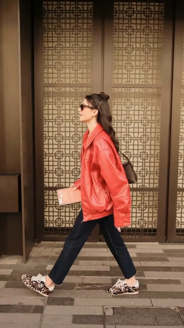 A woman in a red leather jacket and dark jeans walking past a carved wooden screen.